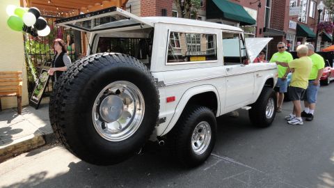1964 Ford Fairlane - White Ford Bronco with a spare tire on the rear, parked on a street with people around.