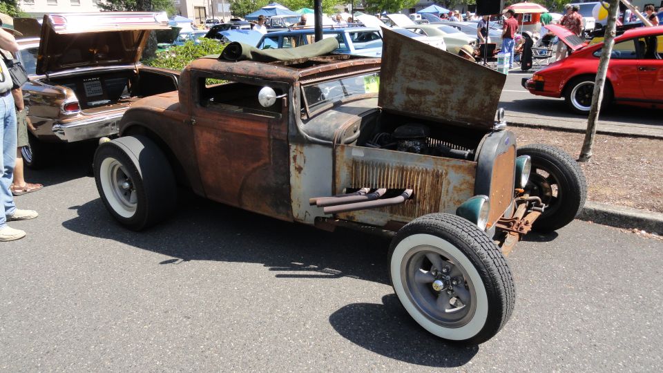 rusted hot rod with open hood and exhaust pipes, parked next to a brown car with open trunk