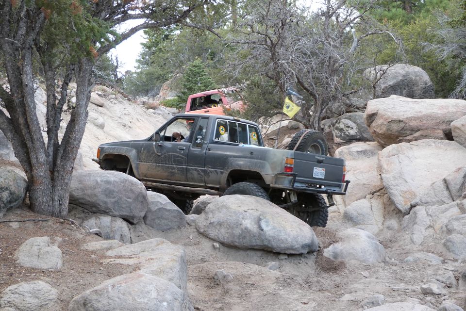 Dark gray Toyota pickup truck on rocky terrain, spare tire mounted on rear, yellow flag on tree