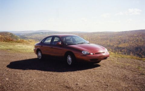 1997 Ford Taurus - A red sedan is parked on a gravel overlook with a scenic view of rolling hills covered in fall foliage.