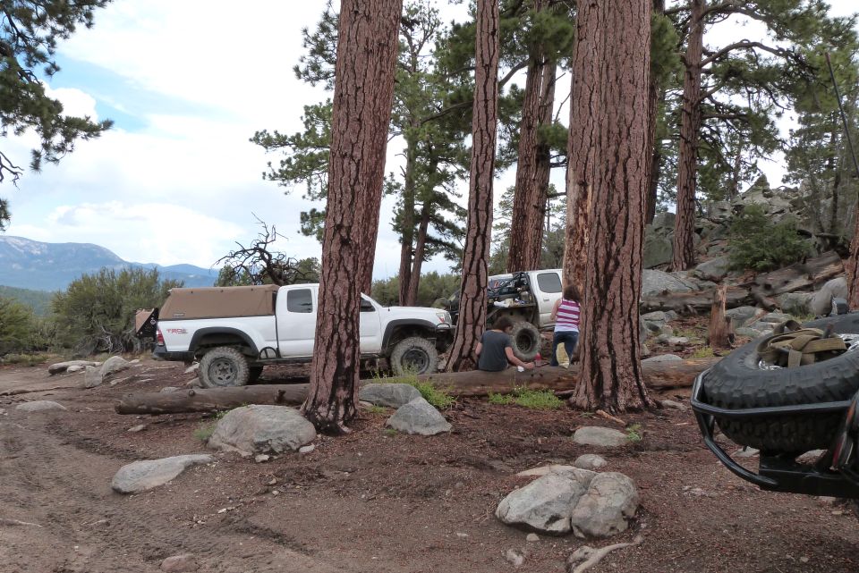 White pickup truck with tan canopy parked on dirt road, trees and rocks around. Another white SUV is visible behind it