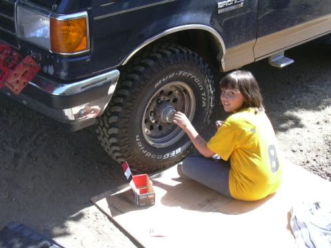 1991 Ford Bronco - A young person in a yellow t-shirt kneels on plywood beside a dark blue Ford Bronco, working on the exposed front hub. The truck runs BFGoo…