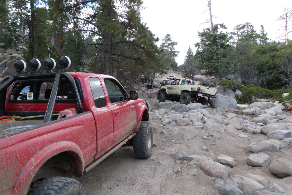 Red pickup truck in foreground, tan off-road vehicle navigating rocks in background