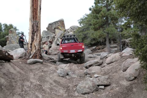 1985 Toyota 4Runner - A red Toyota pickup truck is shown off-roading on a rocky trail, with two people observing the vehicle.
