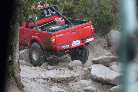 1985 Toyota 4Runner - This red Toyota pickup truck is shown off-roading over a rocky obstacle course, equipped with a spare tire and a red gas can in the bed.