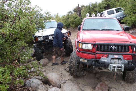 1985 Toyota 4Runner - Two Toyota trucks navigate a challenging off-road trail with large rocks and dense foliage.
