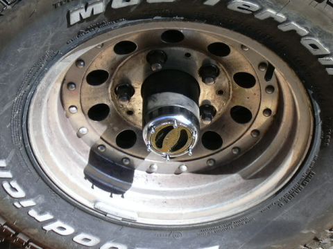 1991 Ford Bronco - Overhead close-up of a BFGoodrich Mud-Terrain tire mounted on a weathered white-spoke steel wheel with a simulated beadlock ring, laid flat…