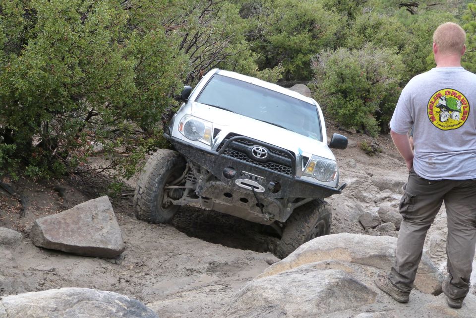 White Toyota Tacoma with aftermarket bumper navigating a rock obstacle, person watching