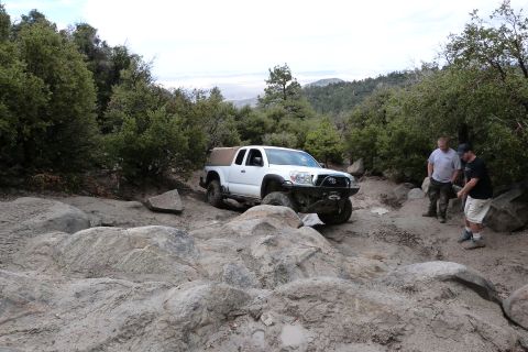 1985 Toyota 4Runner - A white Toyota Tacoma navigates a challenging rocky terrain on a wooded mountain trail with two observers.
