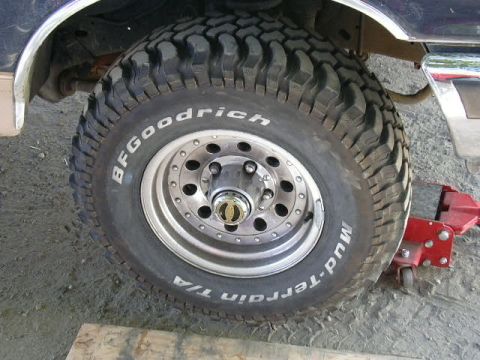 1991 Ford Bronco - Front wheel of a lifted truck photographed from the side on gravel, wearing a BFGoodrich Mud-Terrain T/A white-letter tire on a polished ei…