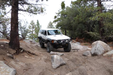 1985 Toyota 4Runner - A white 1985 Toyota 4Runner navigates a challenging off-road course with large rocks and trees.
