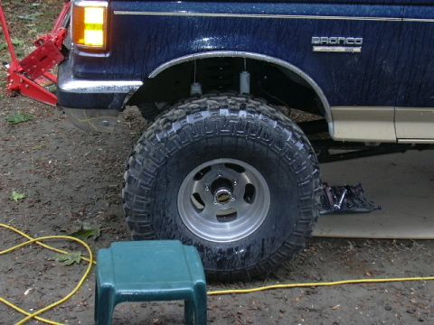 1991 Ford Bronco - Side view of the front corner of a blue and tan two-tone Ford Bronco on an oversized mud-terrain tire and slotted aluminum wheel, with the…
