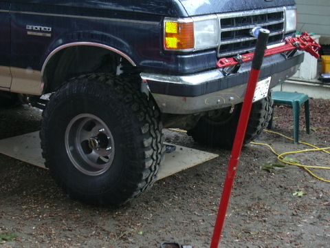 1991 Ford Bronco - Front corner of a dark blue and tan lifted Ford Bronco jacked up on a plywood pad in a gravel driveway, with a red high-lift jack standing…