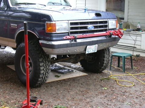1991 Ford Bronco - Blue and tan two-tone Ford Bronco jacked up in a gravel driveway, viewed from a low three-quarter front angle with a red high-lift jack sta…