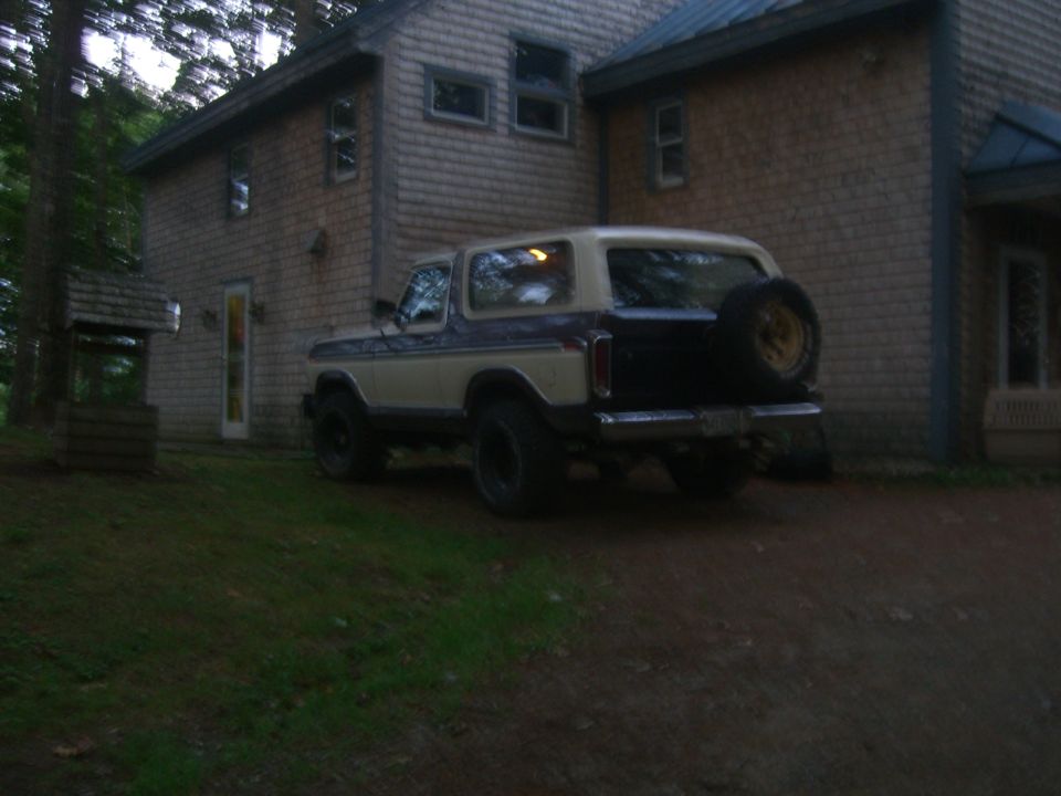 Two-tone beige and dark blue Ford Bronco with a spare tire mounted on the rear, parked on a gravel driveway next to a wooden house