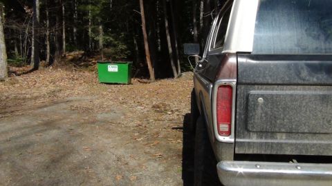 1979 Ford Bronco - The rear of a classic Ford Bronco is visible, parked in a wooded area next to a dumpster.
