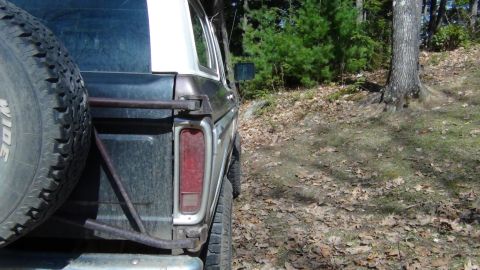 1979 Ford Bronco - A brown and white Ford Bronco is seen from the rear quarter angle, parked outdoors on a wooded dirt road.