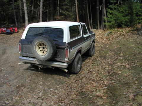 1979 Ford Bronco - A two-tone Ford Bronco sits on a wooded trail, its spare tire prominently displayed on the back.