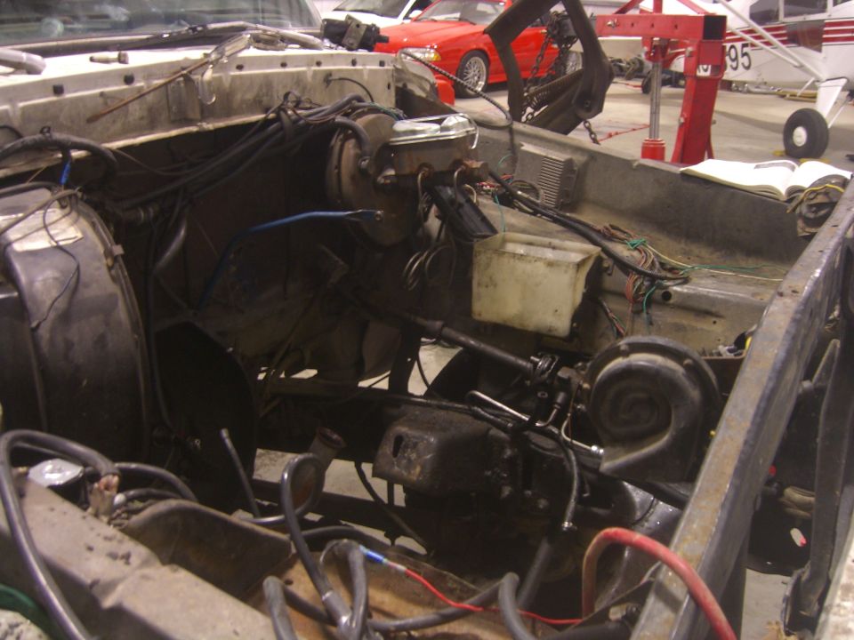 Engine bay of a disassembled vehicle with exposed wires and components. A red car and a small airplane are visible in the background