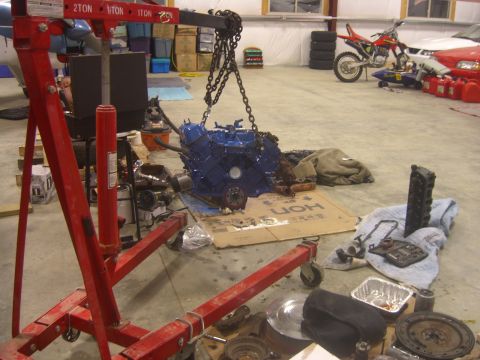 1979 Ford Bronco - Engine hoist lifting a blue engine. Other automotive parts are scattered around the garage floor.