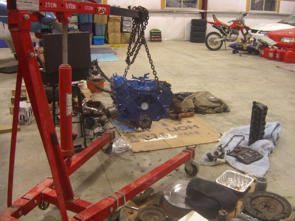 Blue engine suspended from red engine hoist in garage. Parts and tools are on the floor