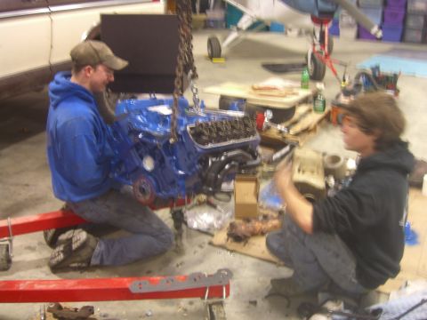 1979 Ford Bronco - Two men attend to a blue engine being hoisted in a workshop, with a small airplane in the background.
