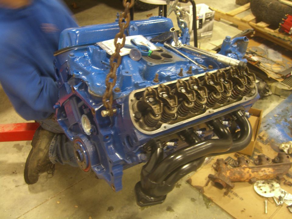 Blue V8 engine with headers being lifted by a chain. Some old exhaust parts are on the floor
