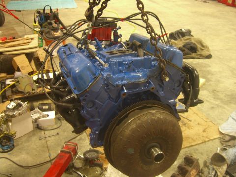 1979 Ford Bronco - A blue V8 engine assembly, complete with a torque converter, is suspended by chains in a workshop environment.