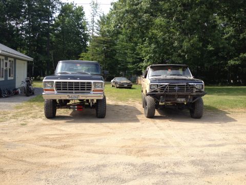1979 Ford Bronco - Two lifted Ford trucks are parked in a dirt lot in front of a building with trees behind them.