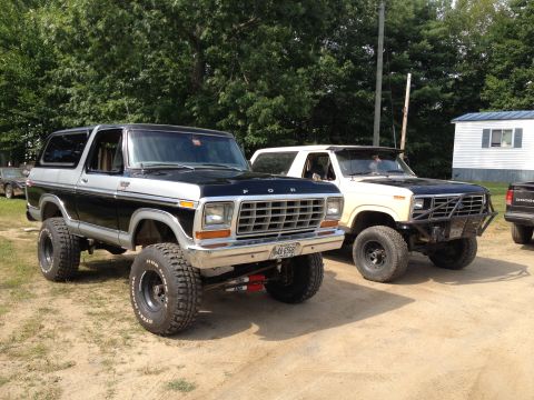1979 Ford Bronco - Two modified Ford Broncos are parked side-by-side on a dirt lot under a sunny sky.