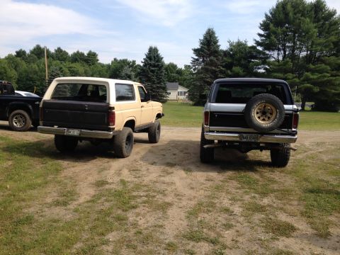 1979 Ford Bronco - Two Broncos are parked in a grassy area with trees and a house in the background.