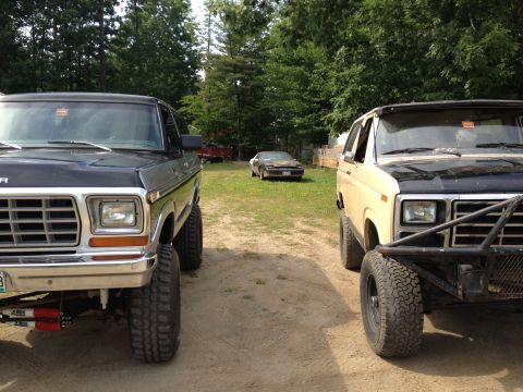 1979 Ford Bronco - Two modified Ford Broncos are parked facing each other on a dirt path, with a car in the distance.