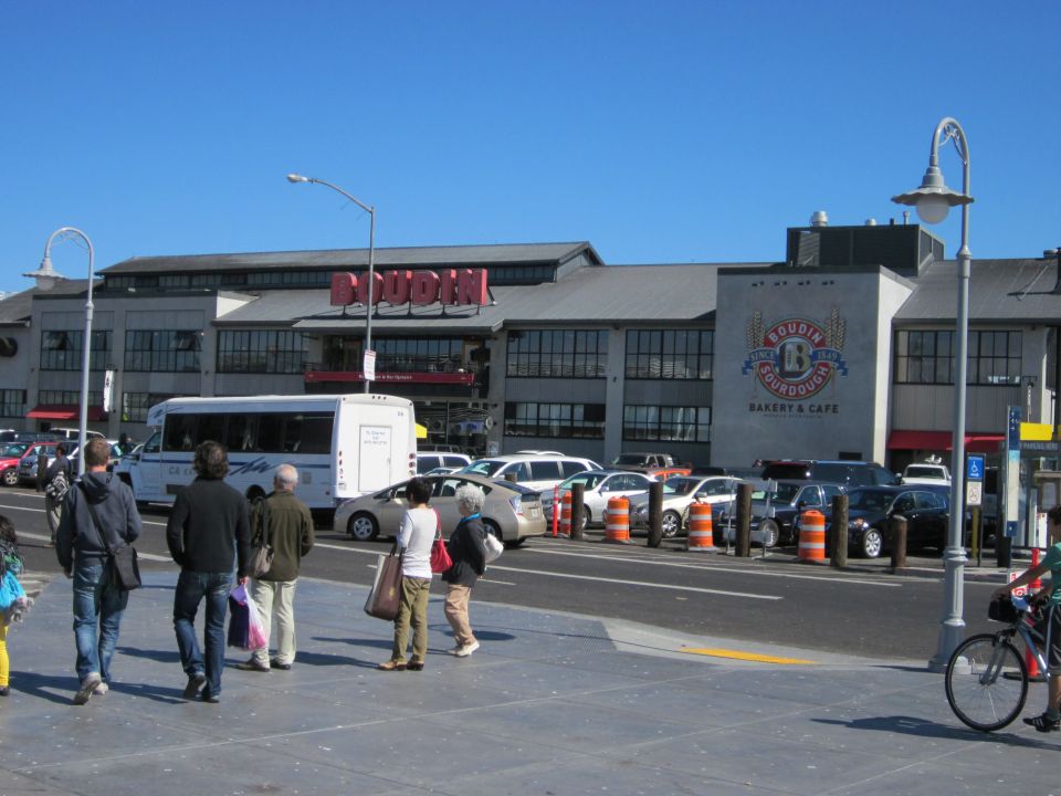 People walking in front of Boudin Bakery with cars and a bus parked nearby