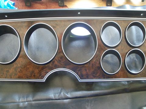 1979 Ford Ranchero - A removed instrument cluster bezel with burl woodgrain applique and bright chrome trim rings, laid face-up on a black vinyl seat. It has th…