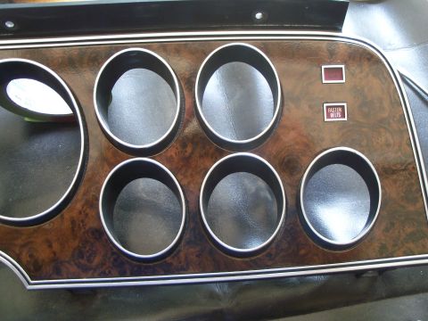 1979 Ford Ranchero - A woodgrain instrument-cluster bezel from a 1970s Ford, removed from the dash and laid on a black seat, with six empty gauge openings trimm…