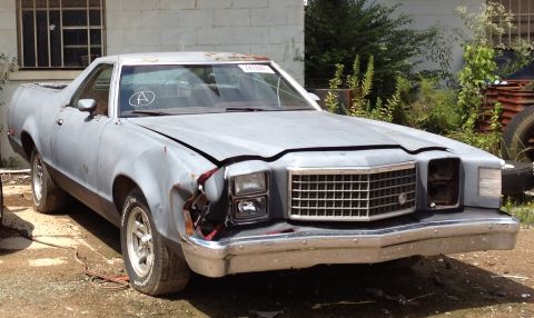 1975 Ford Ranchero - Three-quarter front view of a primer-gray Ford Ranchero coupe utility sitting in a weedy lot beside a cinder-block building, with front-end…