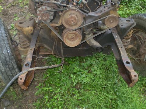 1979 Ford Bronco - Close-up view of engine and front suspension components, including belts and pulleys, resting on grassy ground.