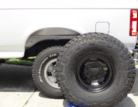 1994 Ford Bronco - Two large off-road tires are shown next to the rear fender of a white SUV. One tire is mounted on a black rim.