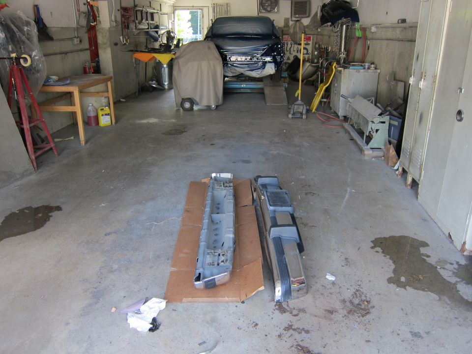 Two car bumpers and a chassis section on a cardboard sheet in a garage