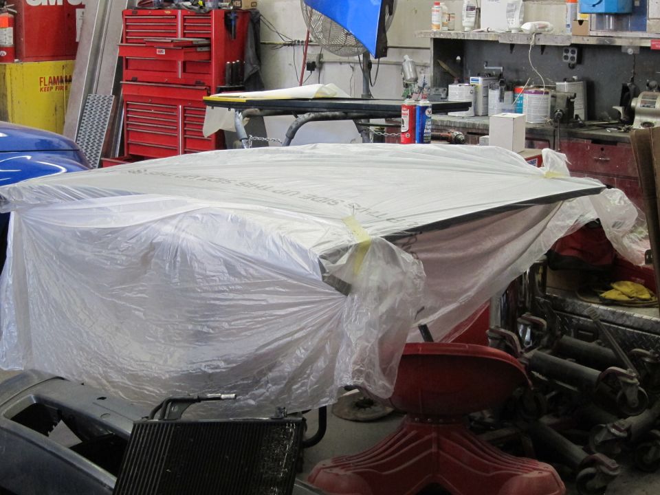 Car hood covered in white plastic sheeting with yellow tape, in a workshop setting
