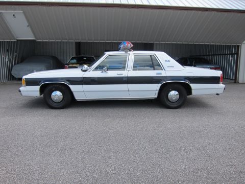 1991 Ford LTD Crown Victoria - A police car is parked in front of a garage with other vehicles inside.