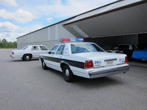1991 Ford LTD Crown Victoria - Two white Ford Crown Victoria police cars are parked outside a large building with an open bay door.