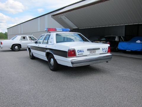 1991 Ford LTD Crown Victoria - A white 1991 Ford Crown Victoria LTD police car is parked outside a hangar, with another similar vehicle visible behind it.