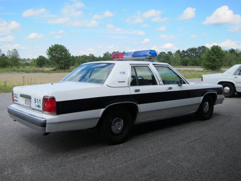 1991 Ford LTD Crown Victoria - A white and black police car is parked on asphalt under a partly cloudy sky.