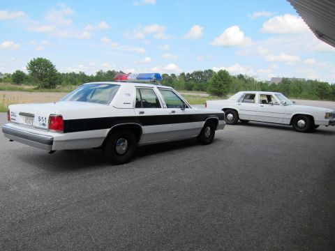 1991 Ford LTD Crown Victoria - Two white police cars are parked on asphalt under a cloudy sky. The foreground car is a 1991 Ford Crown Victoria LTD.