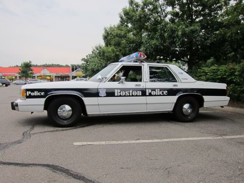 1991 Ford LTD Crown Victoria - A 1991 Ford Crown Victoria LTD police car is seen from the side, parked in a lot with trees and a building in the background.