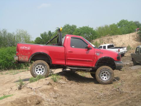 1990 Ford Bronco - A lifted red pickup truck with "4x4 OFF ROAD" decals navigates a dirt track, with other vehicles visible in the background.
