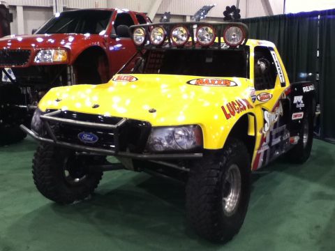 1990 Ford Bronco - A custom yellow off-road truck with racing decals is displayed indoors, with a red truck in the background.