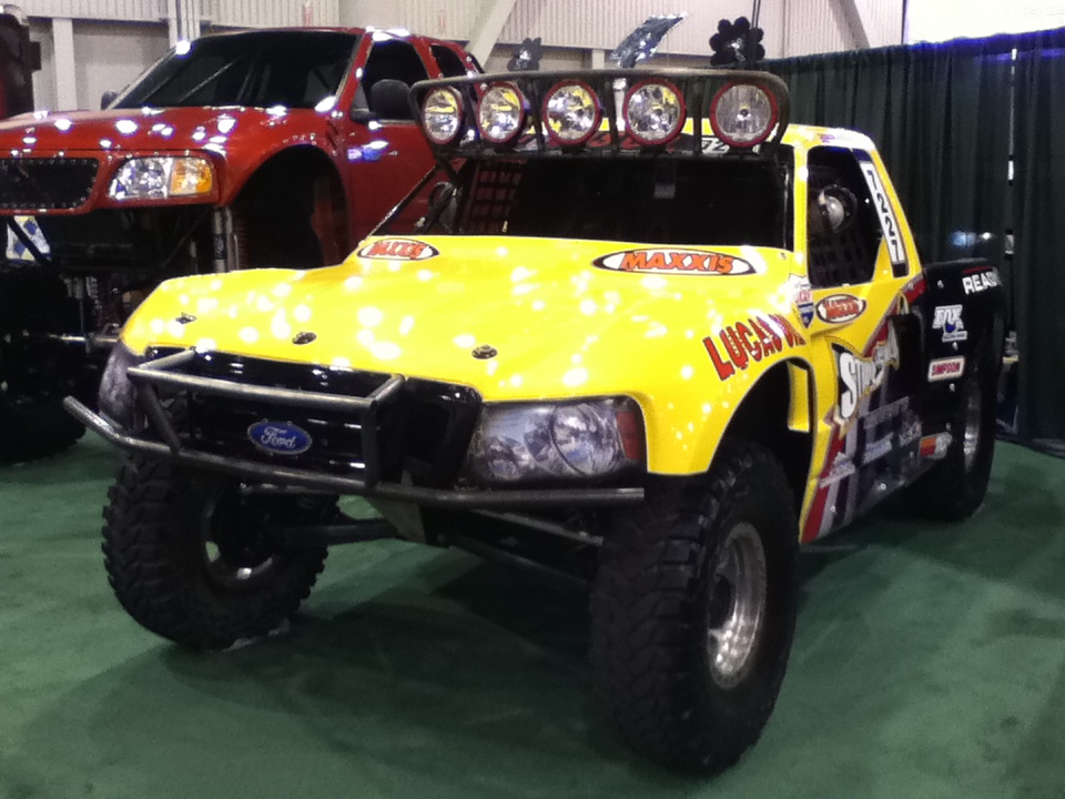 Yellow off-road truck with racing decals, Ford logo on grille. Red truck behind it