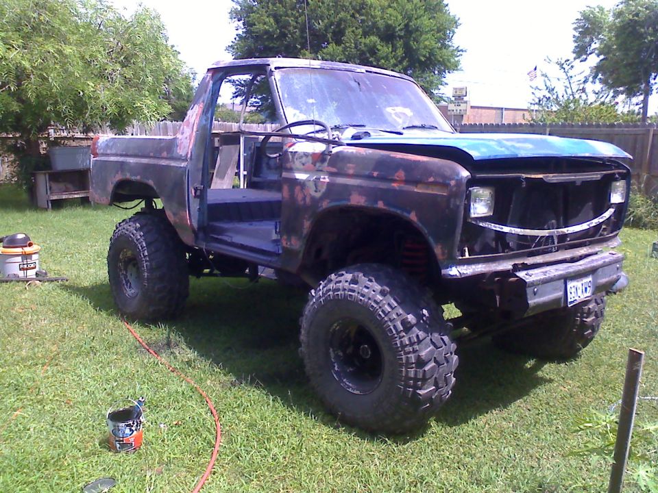 Rusty Ford Bronco with lifted suspension and large tires, doors removed, blue hood